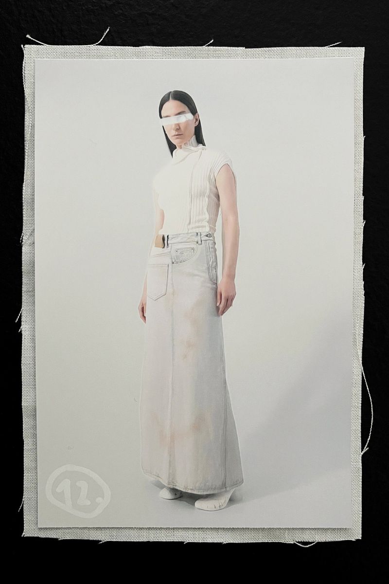 Fashion shot of a model wearing a white top, long skirt, and white visor in front of an off-white background and canvas material.