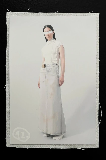 Fashion shot of a model wearing a white top, long skirt, and white visor in front of an off-white background and canvas material.