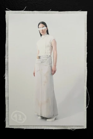 Fashion shot of a model wearing a white top, long skirt, and white visor in front of an off-white background and canvas material.
