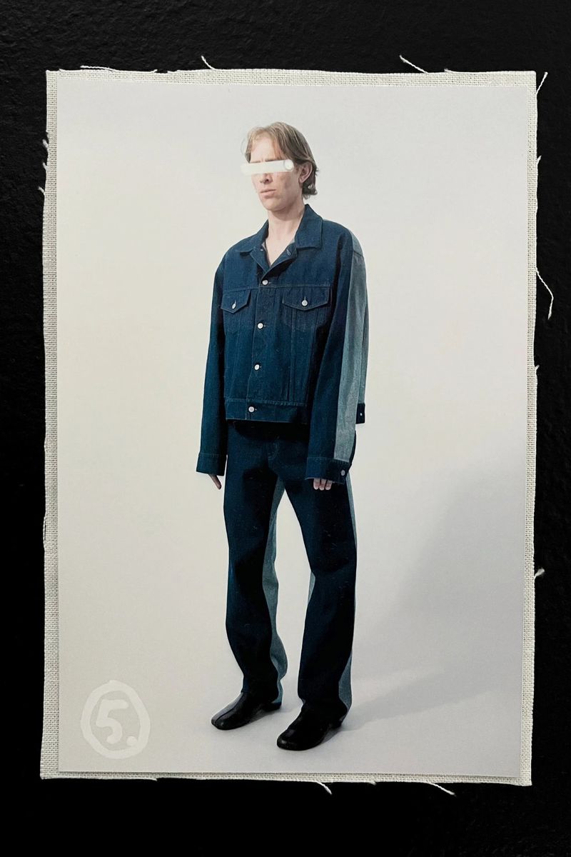 Fashion shot of an adult wearing a denim jacket and pants with partial face covering, standing against an off-white studio backdrop on fabric.