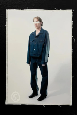 Fashion shot of an adult wearing a denim jacket and pants with partial face covering, standing against an off-white studio backdrop on fabric.