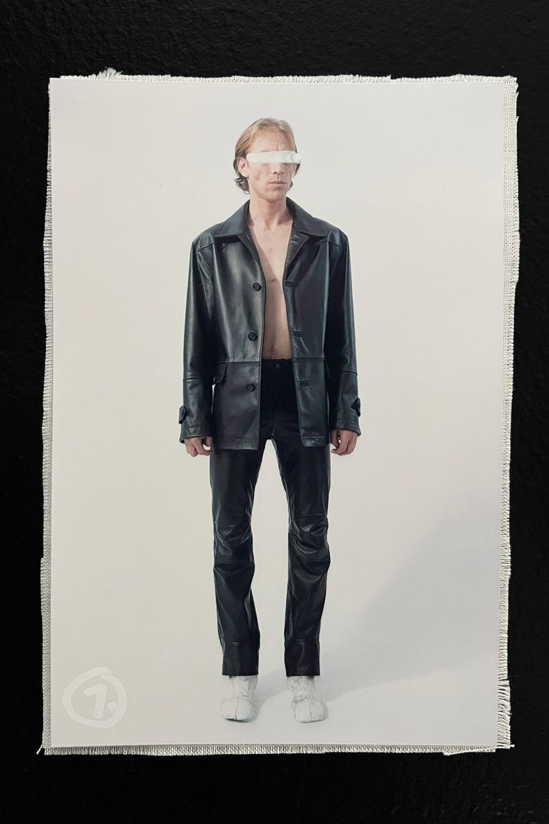 Full-length fashion shot: slender man with blonde hair in a dark leather jacket and pants, eyes obscured by a white bar, against a bright background.