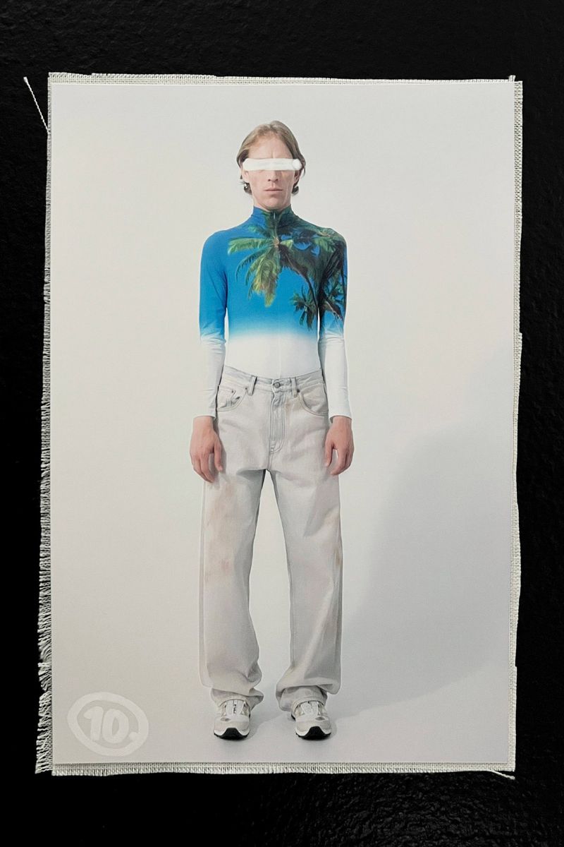 An adult stands in a photo against a white backdrop, wearing a tropical-themed shirt, wide-leg jeans, and white sneakers.