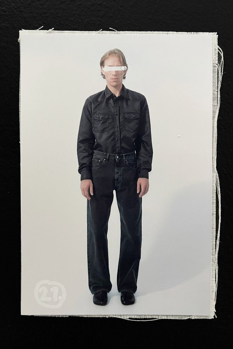 Full-length fashion shot of a man in a black button-down shirt and dark jeans, styled with a white bar obscuring his eyes, against a plain background.