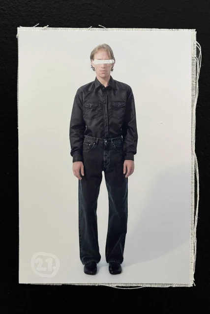 Full-length fashion shot of a man in a black button-down shirt and dark jeans, styled with a white bar obscuring his eyes, against a plain background.