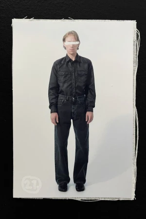 Full-length fashion shot of a man in a black button-down shirt and dark jeans, styled with a white bar obscuring his eyes, against a plain background.