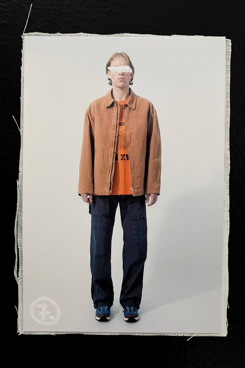Full-body shot of a standing adult wearing a jacket, t-shirt, pants, and sneakers, against a neutral backdrop with a black border.