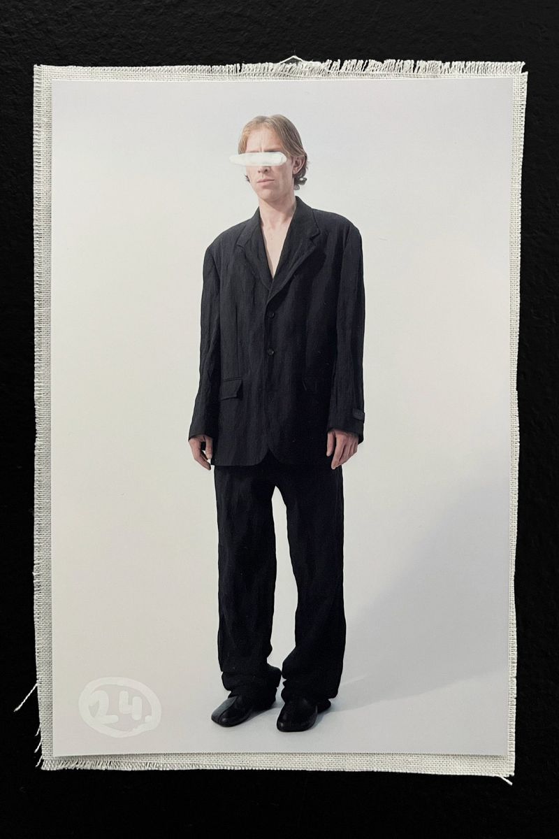 An adult stands in front of a white backdrop wearing a dark suit. A white rectangle covers the eyes. A small white logo is present.