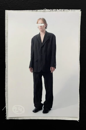 An adult stands in front of a white backdrop wearing a dark suit. A white rectangle covers the eyes. A small white logo is present.