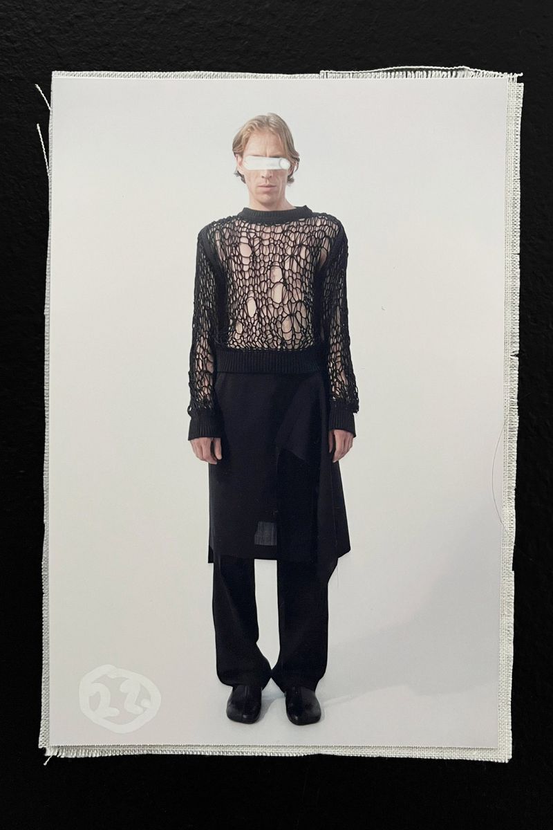Fashion photo of a slender adult wearing a black knit sweater and long skirt-like wrap against a white backdrop on black material.