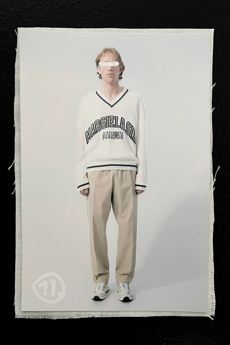 A model with obscured eyes poses in a studio wearing a white v-neck sweater with the text MARGIELA 6:03:48 N 0:18:25 E and beige pants.