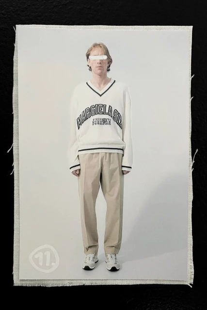 A model with obscured eyes poses in a studio wearing a white v-neck sweater with the text MARGIELA 6:03:48 N 0:18:25 E and beige pants.