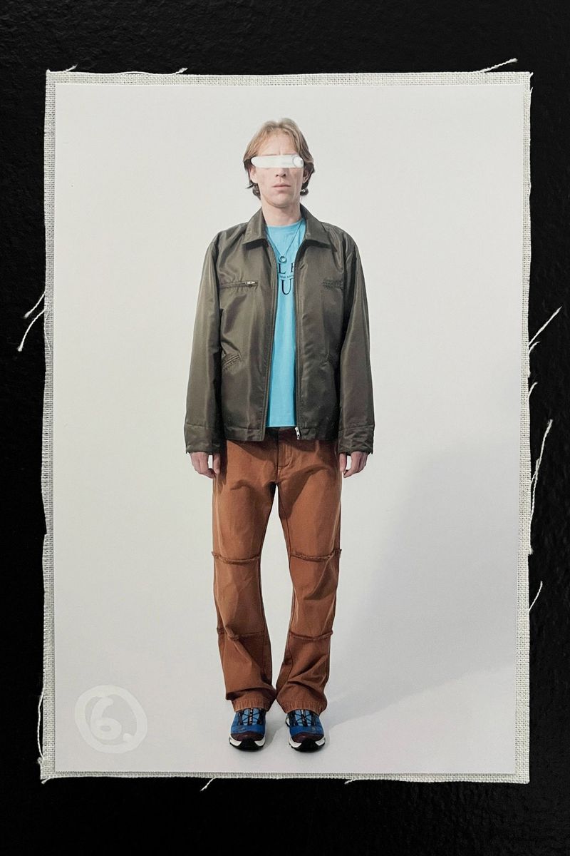 Full-length shot of a person in an olive jacket, blue t-shirt, and copper-colored pants standing against a white backdrop on textured material.