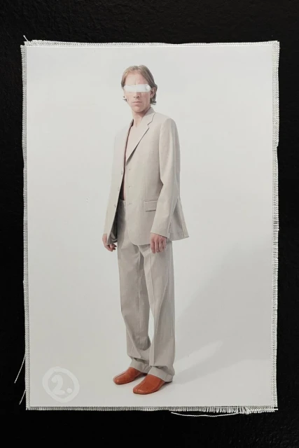 Model in a beige tailored suit and leather shoes with eyes covered by white tape against a neutral studio backdrop.