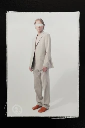 Model in a beige tailored suit and leather shoes with eyes covered by white tape against a neutral studio backdrop.