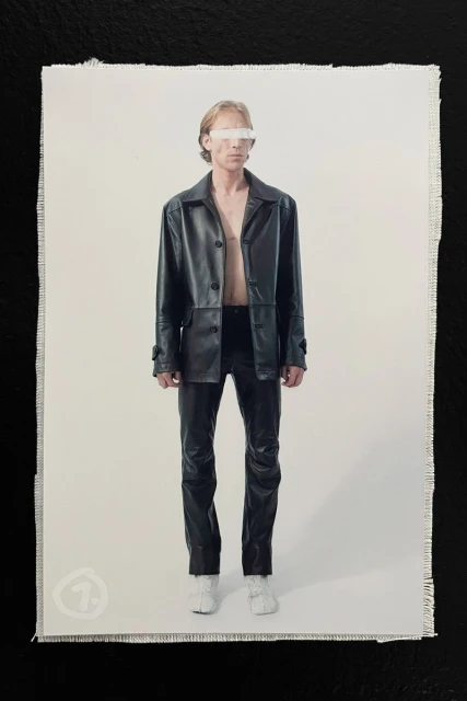 Model wearing a black leather jacket and pants with a white band covering his eyes against a white background.