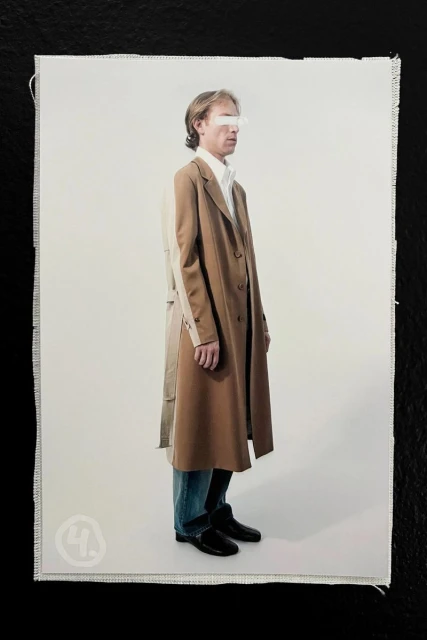 Model wearing a two-tone camel overcoat with blue denim and black shoes against a pale seamless studio background.