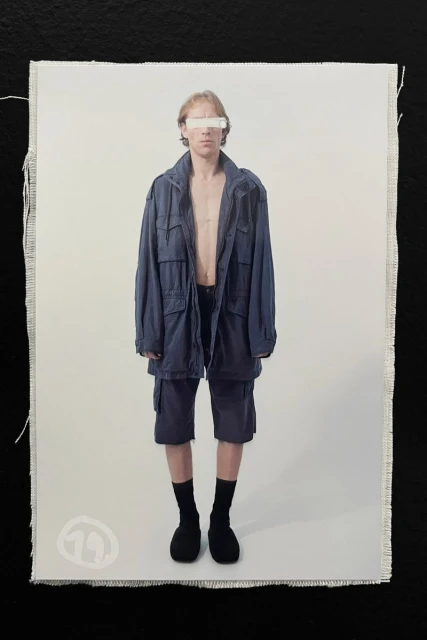 Model wearing a dark blue utility jacket and shorts standing against a light seamless background with eyes redacted.