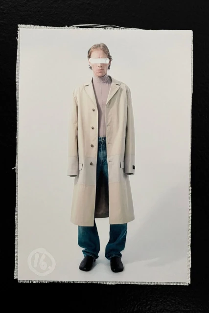 A male model standing wearing a long beige trench coat, turtleneck, relaxed blue jeans, and black shoes against a white backdrop.