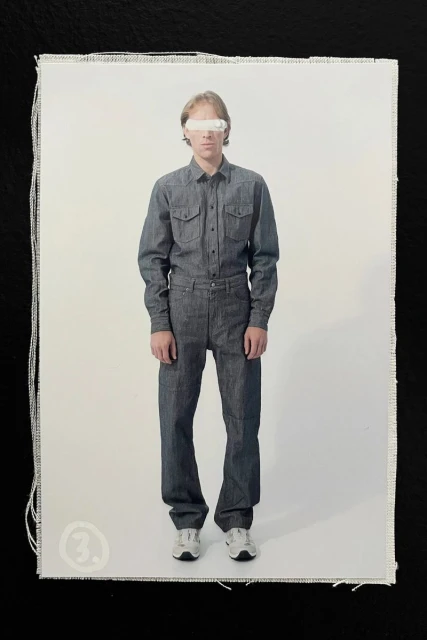 Model wearing a grey denim button-down shirt and matching trousers with a white band covering his eyes against a white studio backdrop.