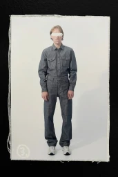 Model wearing a grey denim button-down shirt and matching trousers with a white band covering his eyes against a white studio backdrop.