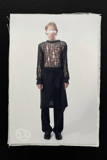 Model wearing a black open-knit mesh top and layered trousers against a white seamless background.