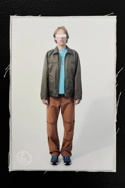 Model wearing olive jacket, blue shirt and brown trousers standing against white seamless background