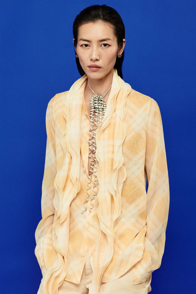 An adult woman poses in front of a blue background, wearing a yellow patterned blouse with ruffles and a large silver necklace.
