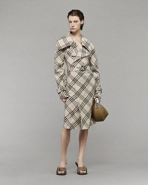 A woman poses in a plaid trench coat dress with a brown handbag and matching sandals, presented against a neutral grey backdrop.