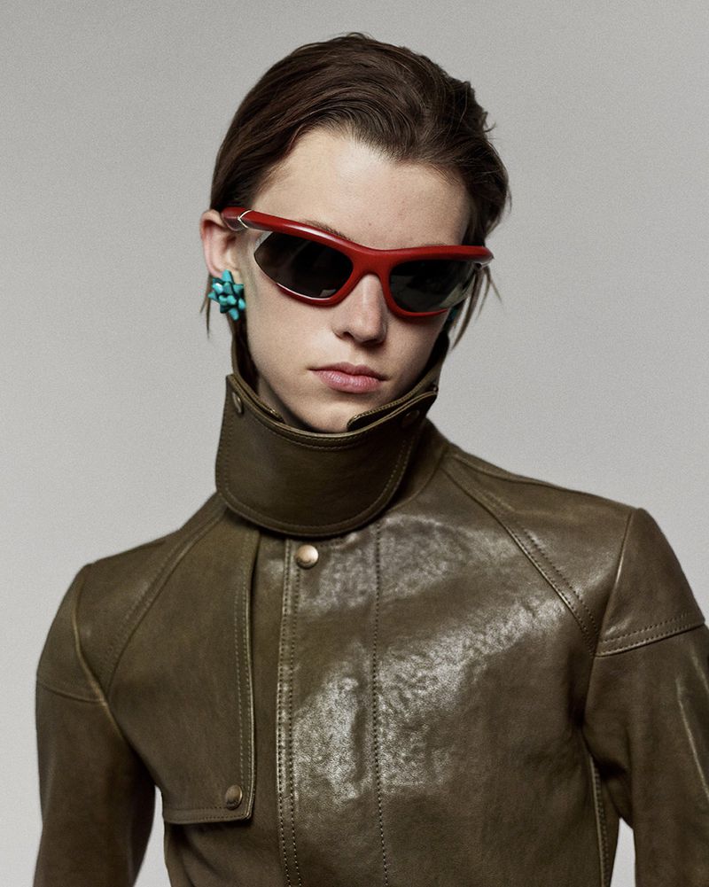 Portrait of a young adult wearing red sunglasses and a brown leather jacket against a neutral background in a studio setting.