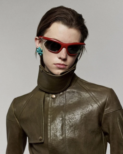 Portrait of a young adult wearing red sunglasses and a brown leather jacket against a neutral background in a studio setting.