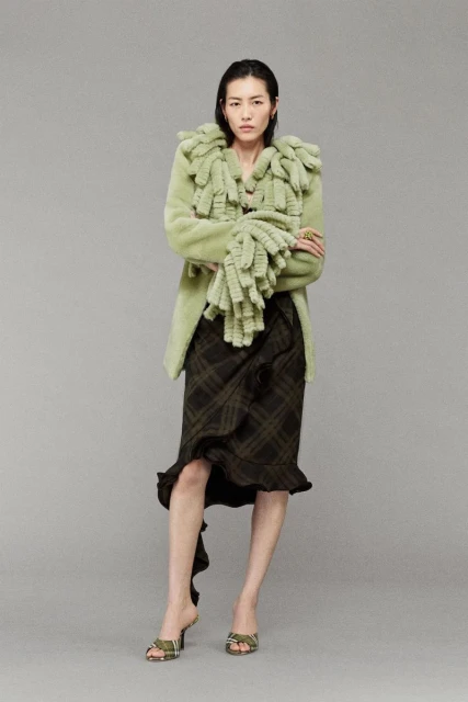 Full-length shot of a slender Asian woman in a light green coat with ruffle details, a plaid skirt, and heeled sandals against gray.