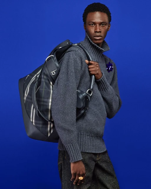 Man poses with large plaid duffel bag in a gray sweater and dark trousers against a bold blue background in a fashion studio shot.