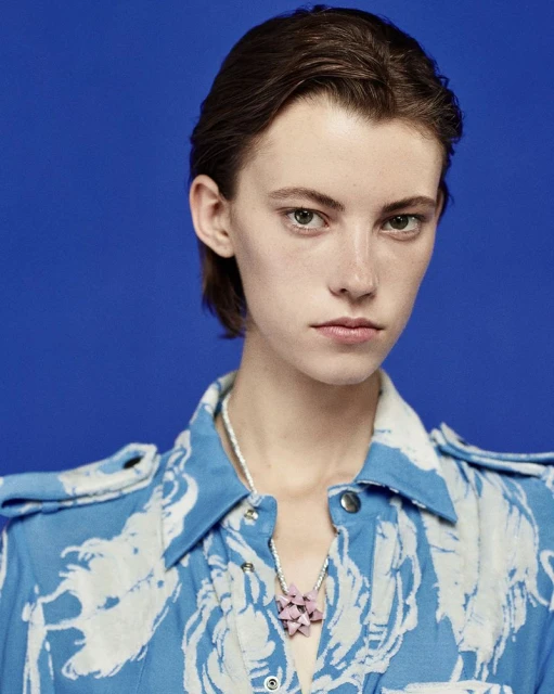 Close-up studio shot of a fair-skinned adult woman with short brown hair wearing a blue patterned shirt and a pale pink geometric necklace.