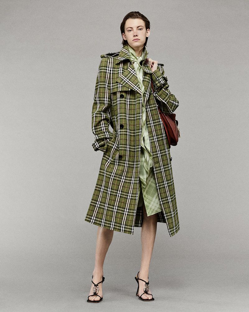 Fashionable adult woman wearing a long olive green plaid trench coat and strappy high-heeled sandals poses in a studio setting.