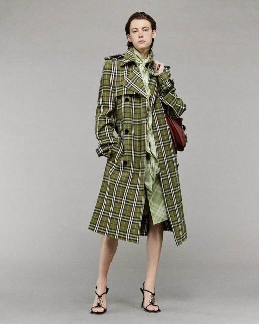 Fashionable adult woman wearing a long olive green plaid trench coat and strappy high-heeled sandals poses in a studio setting.
