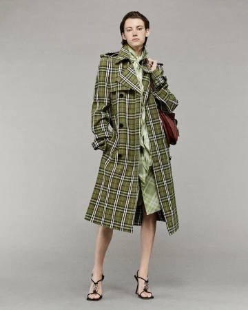 Fashionable adult woman wearing a long olive green plaid trench coat and strappy high-heeled sandals poses in a studio setting.