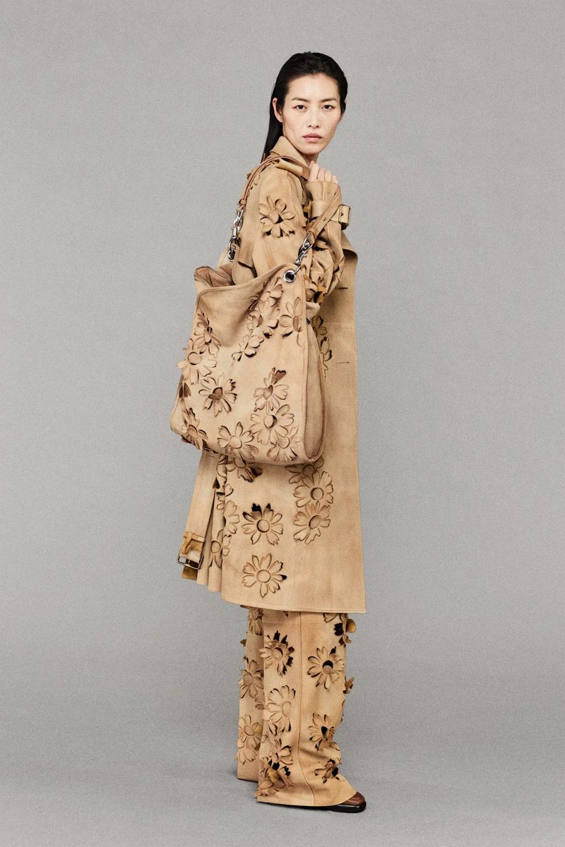 Woman wearing a tan trench coat and pants with laser-cut flower designs, holding a matching tote bag, standing in a neutral studio.