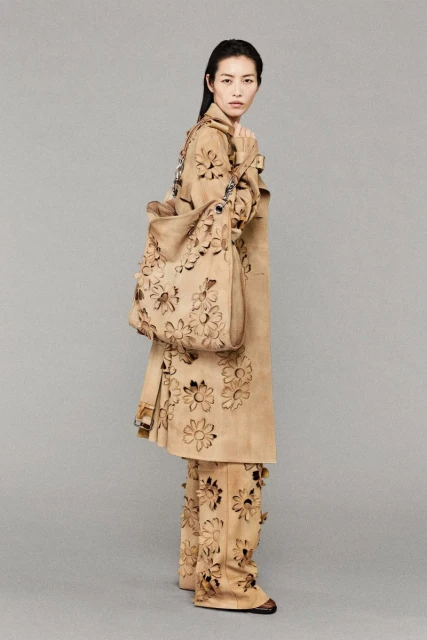 Woman wearing a tan trench coat and pants with laser-cut flower designs, holding a matching tote bag, standing in a neutral studio.