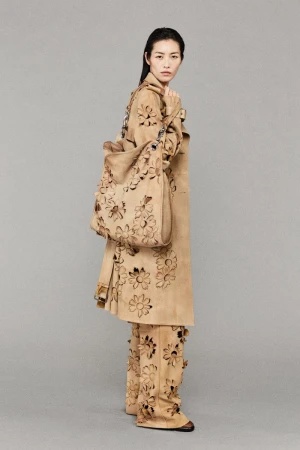 Woman wearing a tan trench coat and pants with laser-cut flower designs, holding a matching tote bag, standing in a neutral studio.