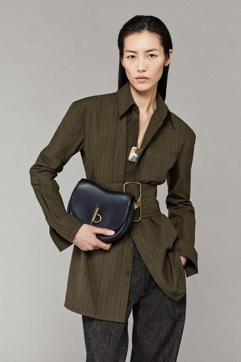 Fashion model wearing a green pinstripe shirt with black purse and grey trousers in a studio setting with a soft focus.