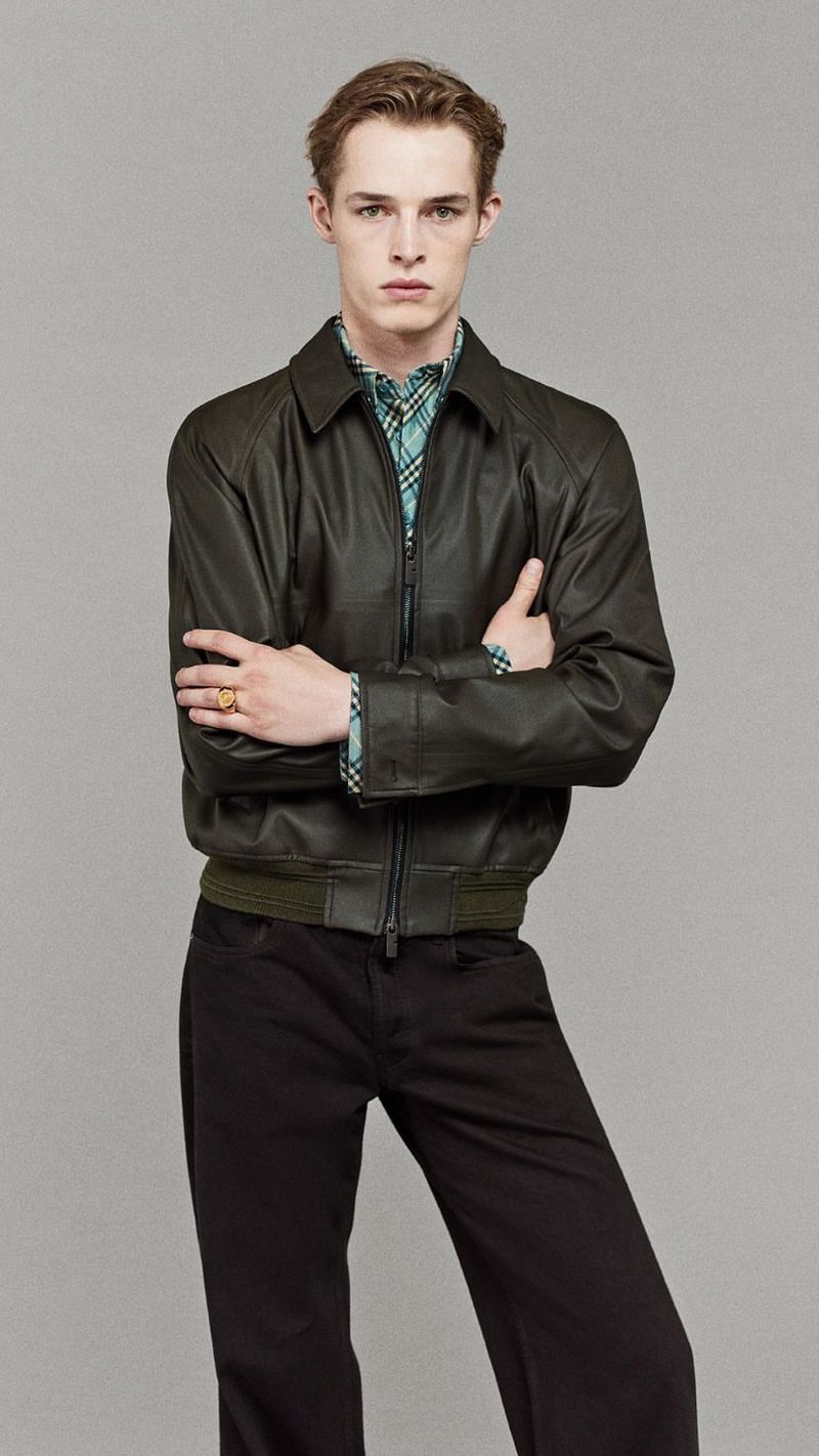 Portrait of a slender adult male in a dark-olive leather jacket and dark pants against a light-grey seamless backdrop in a studio setting.