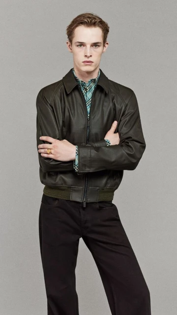 Portrait of a slender adult male in a dark-olive leather jacket and dark pants against a light-grey seamless backdrop in a studio setting.