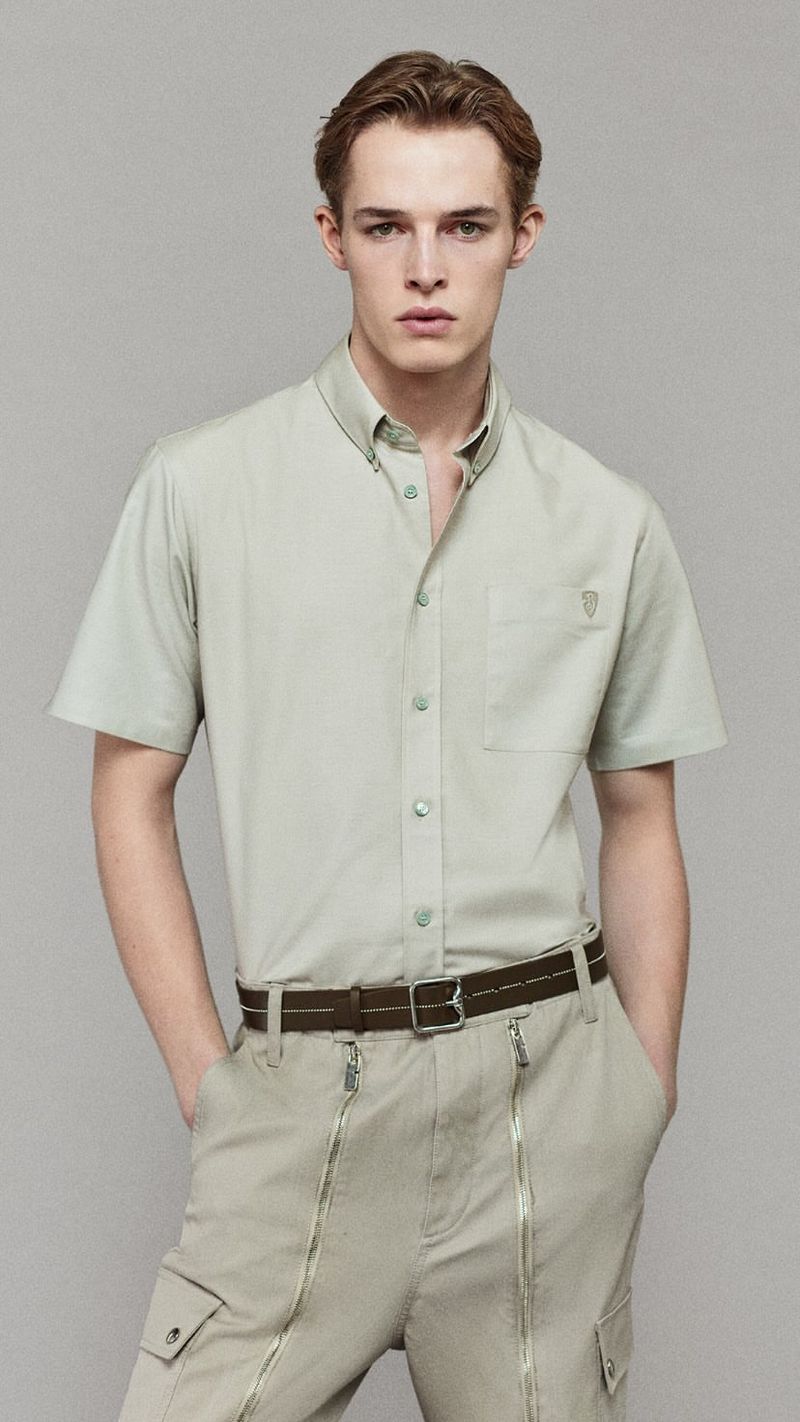 A young adult man stands in a medium shot in a light-green, short-sleeved button shirt with light khaki pants and a brown belt in a studio setting.