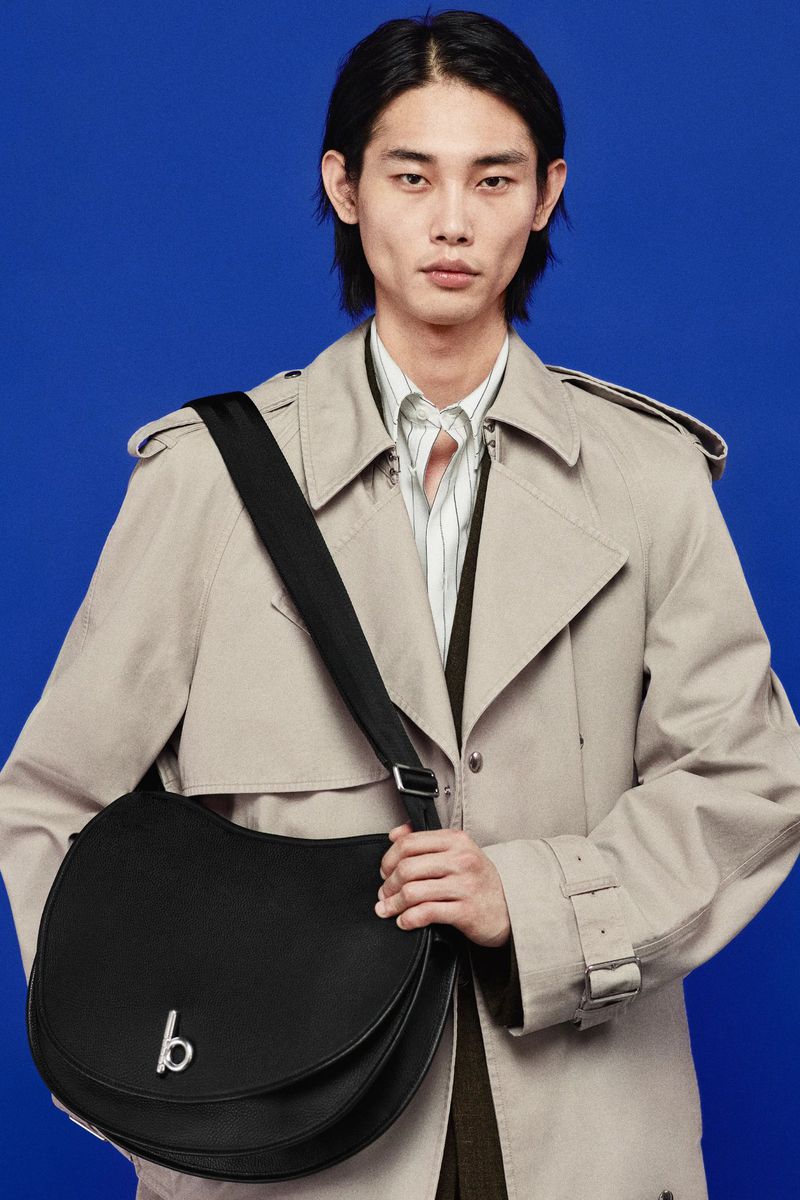 An Asian male model poses in a beige trench coat, wearing a black cross-body bag with silver hardware on a bright blue backdrop.