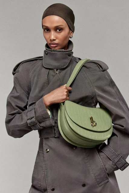 A woman poses in a gray trench coat holding a light green purse, in a studio shot, with neutral gray background. Fashion, style, and elegance.