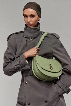 A woman poses in a gray trench coat holding a light green purse, in a studio shot, with neutral gray background. Fashion, style, and elegance.