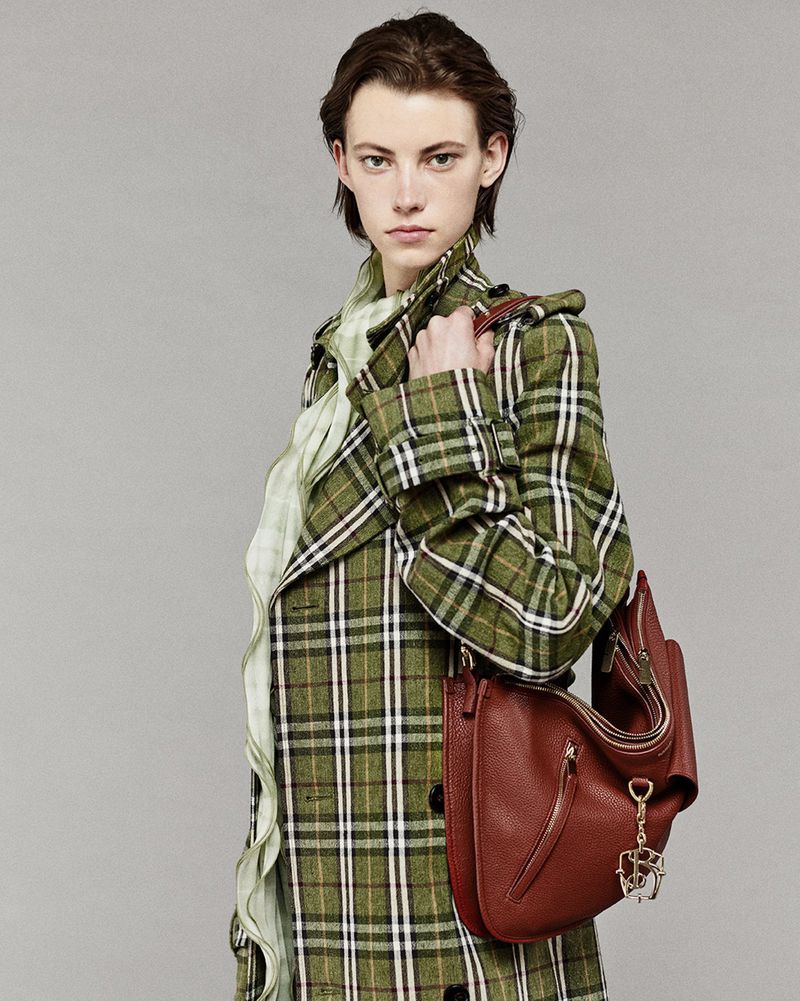 Fashion image of a stylish adult woman wearing a green plaid coat and holding a brown leather shoulder bag in a studio setting.