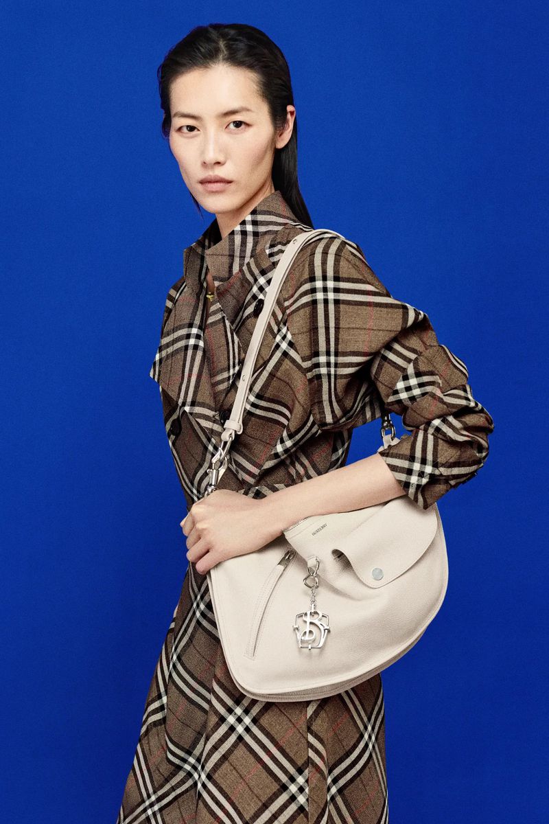 Asian woman poses in plaid dress with ivory satchel against blue background. Fashion shot with medium framing, neutral expression.