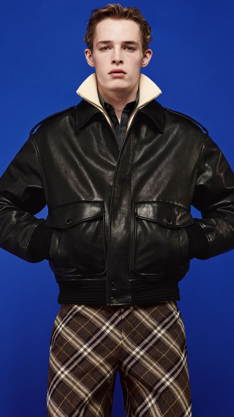 Portrait of a slender adult male model wearing a black bomber jacket and plaid pants against a bright blue backdrop in a studio.